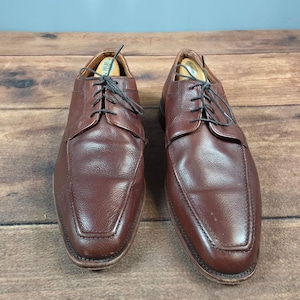 May include: A pair of brown leather dress shoes with a textured finish. The shoes have a classic lace-up design with black laces and a square toe. The shoes are displayed on a wooden surface.