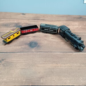 May include: A set of toy train cars, including a black locomotive, a black car with "UNION PACIFIC" text, a red car, and a yellow and brown car. The toy train cars are arranged on a wooden surface.