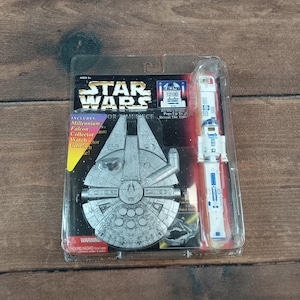 Op de afbeelding: Een Star Wars Millennium Falcon verzamelhorlogeset in een doorzichtige plastic verpakking. De set bevat een Millennium Falcon horlogekast en een R2-D2 horloge. De verpakking toont het Star Wars logo en de tekst "Pops Up To Reveal The Time".