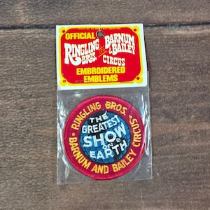 Könnte beinhalten: Ein runder gestickter Aufnäher mit dem Text The Greatest Show On Earth in Weiß und Blau, umgeben von einem roten Rand mit gelbem Text Ringling Bros. and Barnum and Bailey Circus, verpackt in einer durchsichtigen Plastiktüte mit einer Karte.
