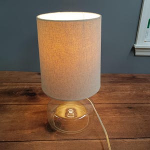 May include: A table lamp with a beige fabric shade and a clear glass base. The lamp is turned on, emitting a warm glow. The lamp is on a wooden surface.