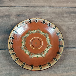 May include: A terracotta-coloured ceramic plate with a decorative design. The plate has a central circular pattern with concentric rings and a border of repeating oval shapes. The design incorporates green, blue, and yellow accents.
