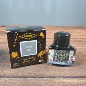 Puede incluir: Un frasco de 40ml de tinta para pluma estilográfica Diamine Silver Fox con tapa negra y base de vidrio cuadrada. La caja de cartón negra incluye gráficos de salpicaduras de tinta amarilla, el logo Diamine y el texto Silver Fox y Made in the U.K.