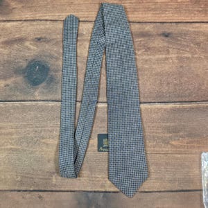 May include: A necktie featuring a repeating grid pattern of small blue squares outlined in gold on a dark background. A black rectangular tag with gold text reading Aquascutum is attached to the fabric near the center of the tie.