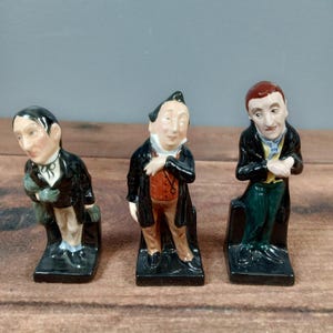 Op de afbeelding: Drie vintage keramische beeldjes van mannen in formele kleding. Elke figuur is beschilderd met gedetailleerde gelaatstrekken en kleding, waaronder zwarte jassen, vesten en broeken. De beeldjes zijn gemonteerd op zwarte rechthoekige bases, mogelijk boekensteunen of decoratieve stukken.