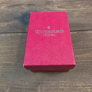May include: A rectangular, textured red box with a lid. The top of the box features the silver-colored text WATERFORD CRYSTAL and a small logo symbol centered above the word Waterford. The box has a structured, solid appearance with sharp edges.