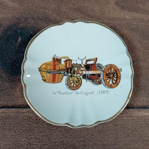 May include: A white, scalloped-edge ceramic plate with a gold rim. The plate features a colorful illustration of a vintage steam-powered vehicle, with the text "le 'Fardier' de Cugnot - 1769".