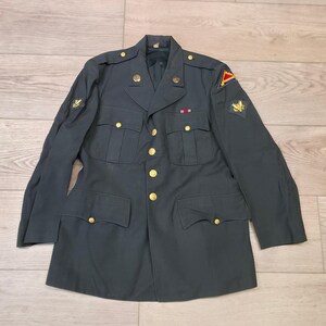 May include: Dark green military jacket with gold buttons and patches. The jacket has four front pockets and a notched collar. Patches are visible on the sleeves. The jacket is laid flat on a light-coloured wooden surface.