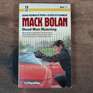 May include: A paperback book cover featuring the title "MACK BOLAN" in large red letters. The cover art depicts a man aiming a gun at a red car. The book is titled "Dead Man Running" and is part of the "Executioner" series.