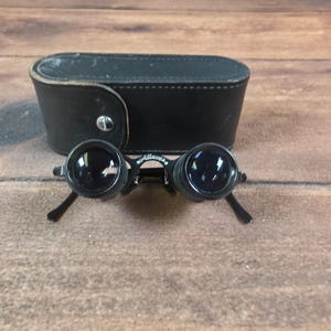 May include: A pair of black Allscope opera glasses with circular lenses and a bridge featuring the brand name in white script. The optical device includes a matching black leather carrying case with a silver-toned metal snap closure button.