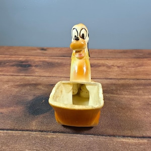 May include: A ceramic planter shaped like a cartoon dog with a tan and cream-colored body. The dog has large white eyes with black pupils, a brown nose, and an open mouth. The planter section is rectangular with rounded edges and a hollow interior.