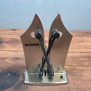 Op de afbeelding: Een zilverkleurige messenslijper met de tekst "Bavarian Edge". De slijper heeft twee schuine messen en zwarte handvatten. De basis is zilverkleurig en heeft een klein logo rechtsonder. Voor keukengebruik.