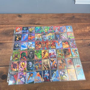 Puede incluir: Una colección de cromos coloridos con varios personajes de cómics. Las cartas están en fundas protectoras y dispuestas sobre una superficie de madera. Las cartas muestran ilustraciones vibrantes y nombres de personajes.