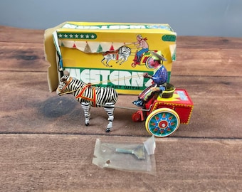 May include: A vintage tin wind-up toy featuring a zebra pulling a red cart with a driver figure. The toy includes a metal key in a plastic bag and a yellow cardboard box labeled WESTERN with a graphic illustration of the toy on the front.
