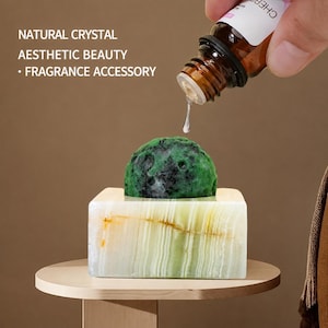 Puede incluir: Una piedra de cristal verde redonda y texturizada descansa sobre una base rectangular de piedra translúcida con bandas blancas y verde pálido. Una gota de líquido transparente cae de un frasco cuentagotas de vidrio marrón. Texto: NATURAL CRYSTAL AESTHETIC BEAUTY • FRAGRANCE ACCESSORY.