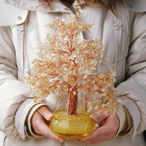 May include: A decorative tree sculpture with a copper wire trunk and branches, adorned with yellow-orange gemstone leaves. The tree sits in a yellow glass pot with the words "Fortune & Wealth" etched on the side. The tree is held in front of a white jacket.