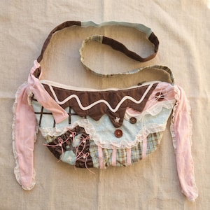 May include: A handmade shoulder bag with a distinctive design. The bag features a brown scalloped top, light blue and plaid fabric, and pink accents. It has a long strap and decorative buttons and lace. The bag is a mix of textures and colours.