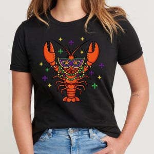 Può includere: T-shirt nero con un design a forma di aragosta a fumetti. L'aragosta arancione indossa una maschera e perline di Mardi Gras, con simboli del giglio. Il design include accenti gialli e verdi, creando una grafica festosa.