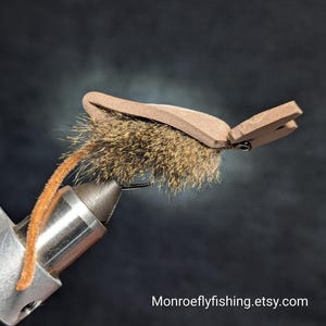 May include: A close-up of a handmade fly fishing lure. The lure features a brown foam body, a tuft of brown fur, and a curved hook. An orange, fuzzy tail extends from the lure. The text "Monroeflyfishing.etsy.com" is visible.