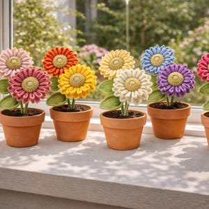 May include: A row of colorful crochet flowers in small terracotta pots. The flowers are in shades of pink, orange, yellow, white, blue, and purple, with green leaves. The pots are arranged on a white windowsill.