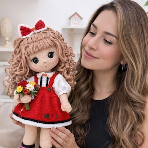Może przedstawiać: Ręcznie robiona szydełkowa lalka z kręconymi brązowymi włosami, czerwoną sukienką i białą bluzką. Lalka ma czerwoną kokardę i trzyma mały bukiet kwiatów. Oczy lalki są duże i czarne. Lalka jest uroczym przykładem rękodzieła.