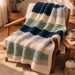 May include: A handmade, striped crochet blanket in shades of navy blue, green, gray, and cream. The blanket is draped over a wooden armchair, showcasing its textured stitch pattern. The cozy throw is a perfect addition to any living space.