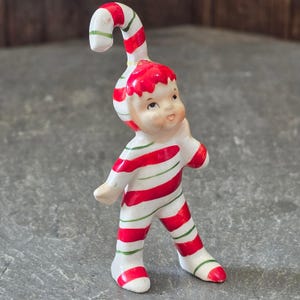 Puede incluir: Una figura de cerámica de un pequeño personaje con un traje de rayas blancas, rojas y verdes y un gorro a juego con la parte superior curvada como un bastón de caramelo. La figura tiene la cara pintada con ojos oscuros y sonriente.