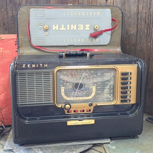 May include: A vintage Zenith Trans-Oceanic portable radio in a black case with a gold-toned control panel and frequency dial. The lid features a detachable grey Wavemagnet antenna with a red strap. The front displays the Zenith brand name.