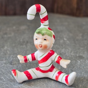 May include: A ceramic figurine of a seated child wearing a white suit with red and green diagonal stripes. The figure has a candy cane shaped hat extension, green hair, and a painted face with rosy cheeks and red lips, finished in a glossy glaze.