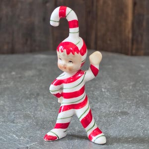 Puede incluir: Una figura de cerámica de un pequeño personaje con un traje de rayas rojas y blancas tipo bastón de caramelo. El personaje lleva un gorro a juego que se curva en la parte superior, con finas líneas verdes que acentúan las rayas rojas del diseño.