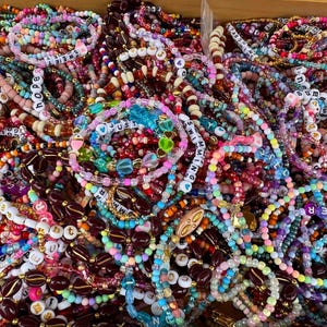 Op de afbeelding: Een verzameling kleurrijke kralenarmbanden met diverse ontwerpen en tekst. De armbanden hebben een mix van kralen in verschillende kleuren, vormen en maten. Sommige armbanden hebben letterkralen met woorden als "hoop" en "zomer".
