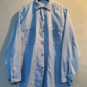 May include: Pale blue denim shirt with long sleeves, two chest pockets, and button fastenings. The shirt is hanging on a wooden hanger against a neutral wall. The shirt has a classic collar and buttoned cuffs.