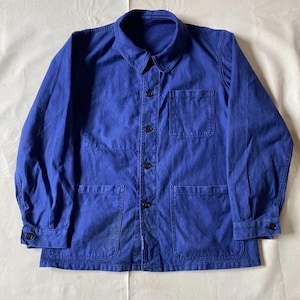 May include: A solid royal blue chore coat featuring a classic fold-over collar, long sleeves with buttoned cuffs, and a front button-down closure. The jacket includes one chest pocket on the left side and two large patch pockets at the waist.