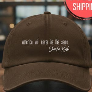 以下が含まれることがあります： カーブしたつばが付いた茶色の野球帽。帽子には白い文字で「America will never be the same. Charlie Kirk」と書かれています。帽子は木製の表面に表示されています。