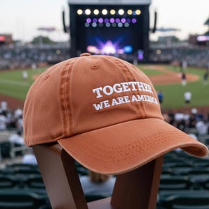 May include: A burnt orange baseball cap with the text "TOGETHER WE ARE AMERICA" in white. The cap is displayed on a wooden stand, with a baseball field and stadium in the background.