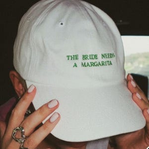 Op de afbeelding: Witte baseballpet met de tekst "THE BRIDE NEEDS A MARGARITA" in groene letters. De pet heeft een gebogen klep en een knoop aan de bovenkant. De pet wordt door een persoon vastgehouden.