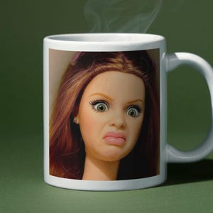 Pode incluir: Caneca de cerâmica branca com o rosto de uma boneca com expressão de surpresa. A boneca tem cabelo castanho avermelhado, olhos verdes e boca franzida. Vapor sobe do topo da caneca, sugerindo uma bebida quente.