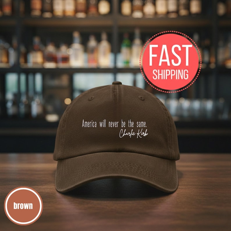 以下が含まれることがあります： カーブしたつばが付いた茶色の野球帽。帽子には白い文字で「America will never be the same. Charlie Kirk」と書かれています。帽子は木製の表面に表示されています。