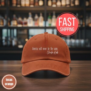 以下が含まれることがあります： 「America will never be the same. Charlie Kirk」の白い文字が入ったテキサスオレンジの野球帽。木製の表面に帽子が表示されています。