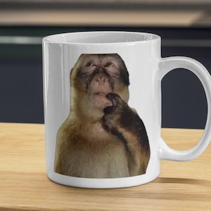 Peut inclure: Mug en céramique blanche avec un mème d'un singe pensif, le doigt sur la bouche. Le singe a une fourrure brune et se détache sur un fond blanc. La tasse a une anse incurvée.