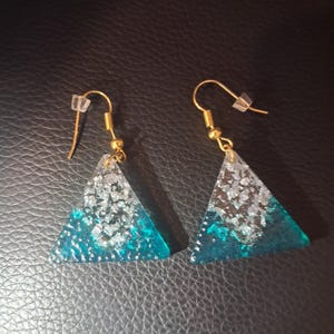 Puede incluir: Un par de pendientes dorados con colgantes triangulares. Los colgantes presentan un diseño turquesa y plateado, con un gancho transparente para la oreja.