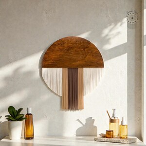 Puede incluir: Un adorno de pared de madera con una parte superior semicircular y una franja de fibras blancas, beige y marrones. El arte de la pared está montado en una pared de color claro. La imagen también muestra una pequeña planta en maceta y varias botellas de color ámbar.