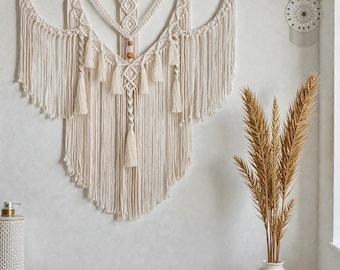 Puede incluir: Un gran tapiz de macramé color crema con una barra de madera. El diseño presenta intrincados nudos, borlas y un patrón geométrico. La decoración de la pared es de estilo bohemio y añade un toque de textura a cualquier habitación.
