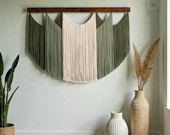 Puede incluir: Un gran tapiz de macramé con una barra de madera. El diseño presenta una sección central de color crema flanqueada por secciones verde oliva. La decoración de la pared se exhibe sobre un suelo de madera clara.
