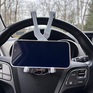 May include: A gray smartphone car mount attached to a black steering wheel. The mount holds a smartphone with a black screen. The background shows bare trees and a cloudy sky.