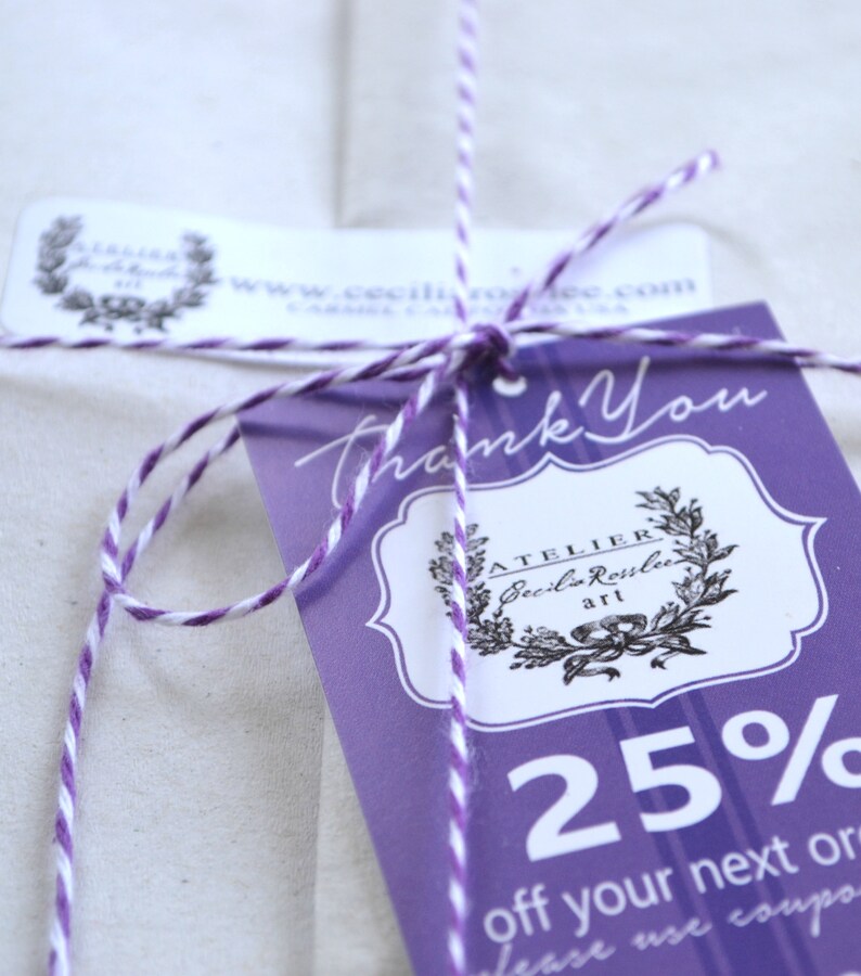 May include: A close-up of a gift tag tied with purple and white twine. The tag is purple with white text that says "Thank You" and features a logo and the text "ATELIER Cecilia Rosslee art". The tag also includes the text "25% off your next order".