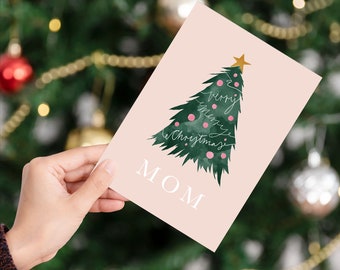 May include: A pink Christmas card with a watercolor Christmas tree design. The tree is green with pink ornaments and a gold star. The words "very merry Christmas" are written on the tree. The word "MOM" is printed in white at the bottom.