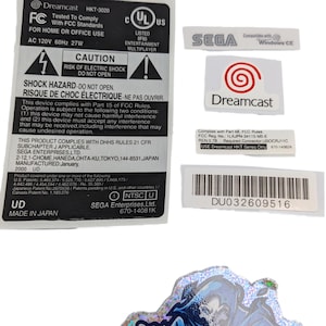 Può includere: Una collezione di etichette e adesivi, tra cui un'etichetta Dreamcast, un'etichetta Sega e un adesivo olografico Gamer Ghost. Le etichette contengono testo come "QC FAIL" e "Made in Japan."