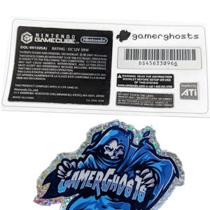 Puede incluir: Una pegatina holográfica con un personaje azul de la Parca y el texto "GAMERGHOSTS". La pegatina tiene un borde brillante. También es visible una etiqueta de Nintendo GameCube.