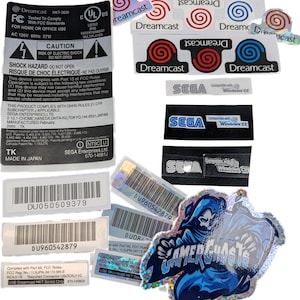 May include: A collection of Sega Dreamcast labels and stickers. The labels feature the Dreamcast logo in red and blue, along with Sega branding. Some stickers have a holographic finish, including a Gamer Ghosts design.